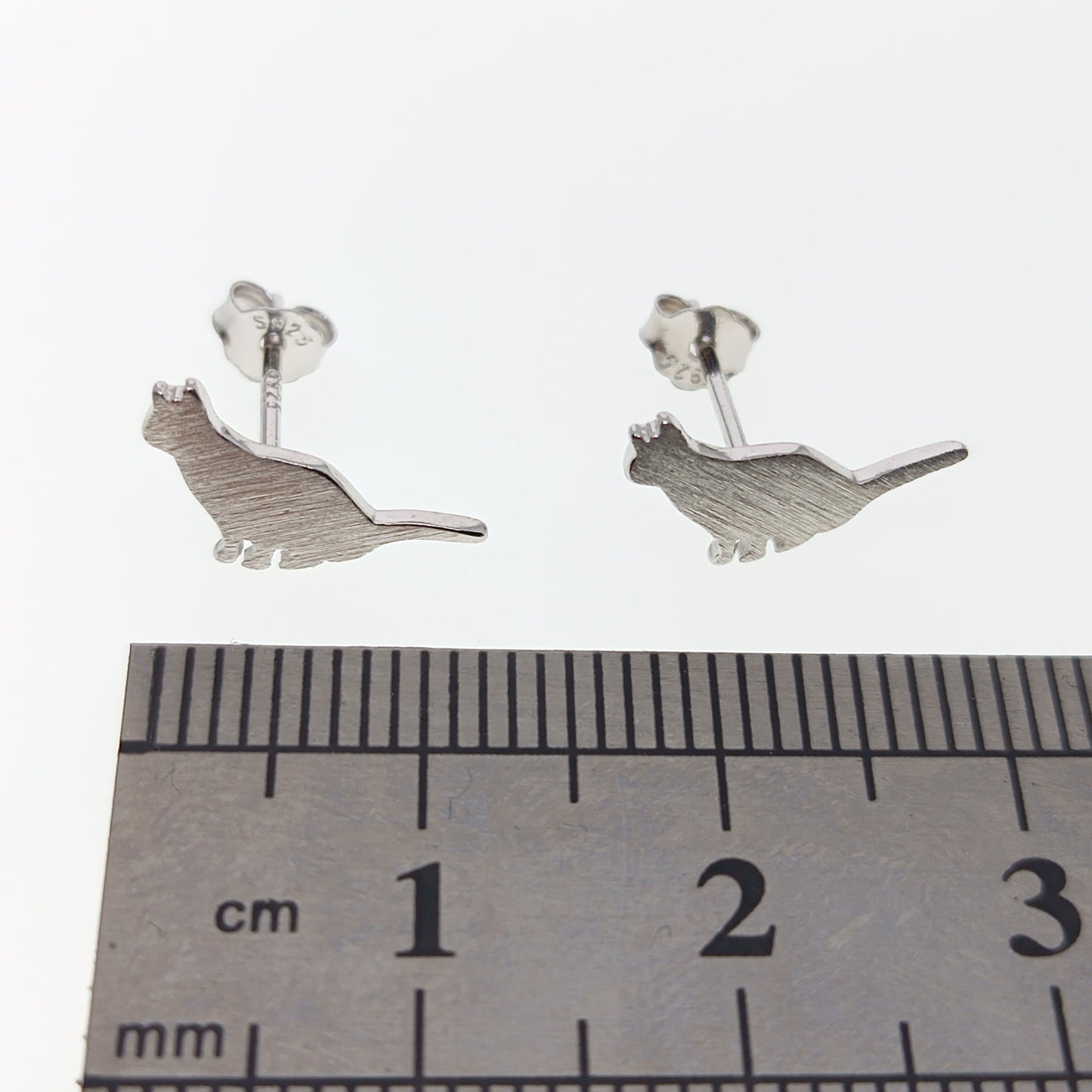 Silver cat-shaped earrings on a white background with a ruler for scale.