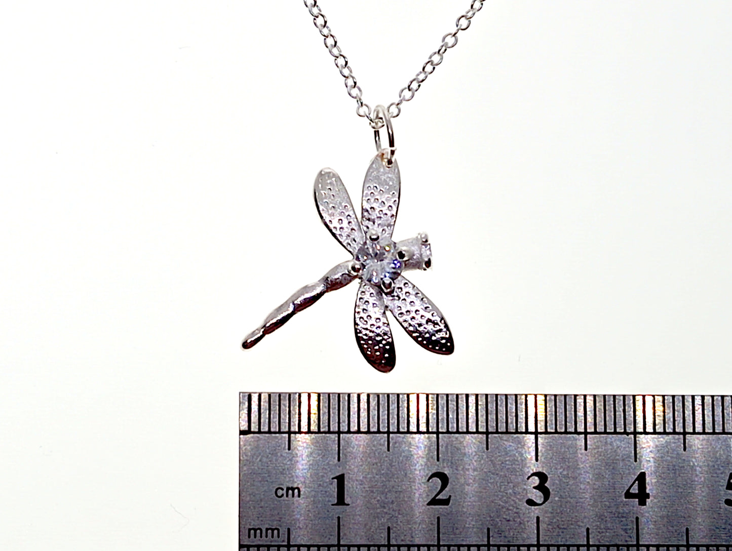 Silver dragonfly pendant on a chain with a ruler for scale on a white background