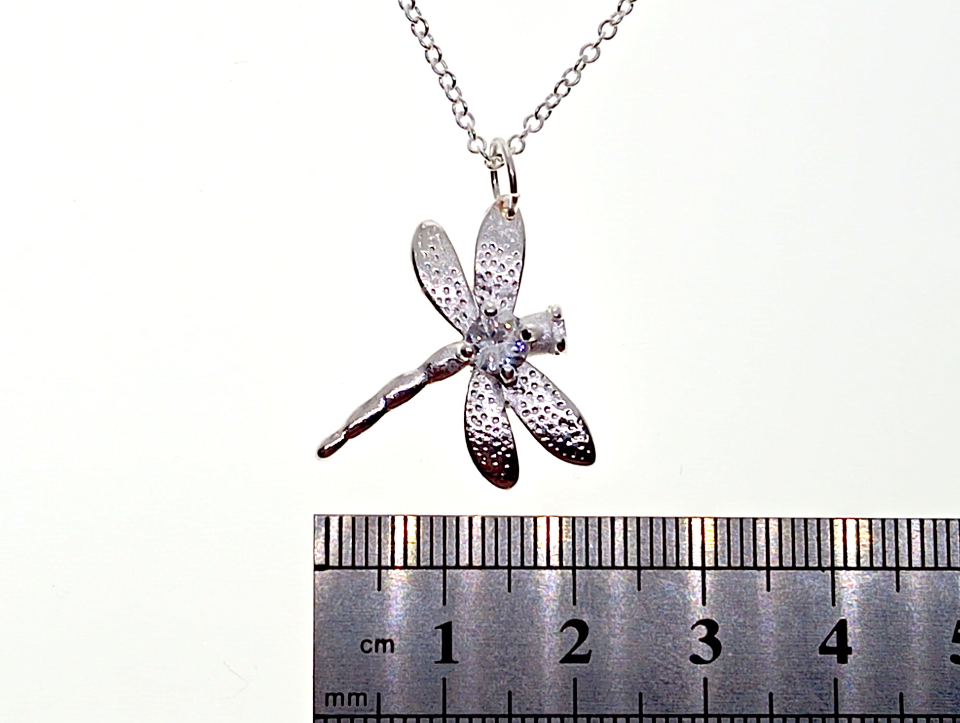 Silver dragonfly pendant on a chain with a ruler for scale on a white background